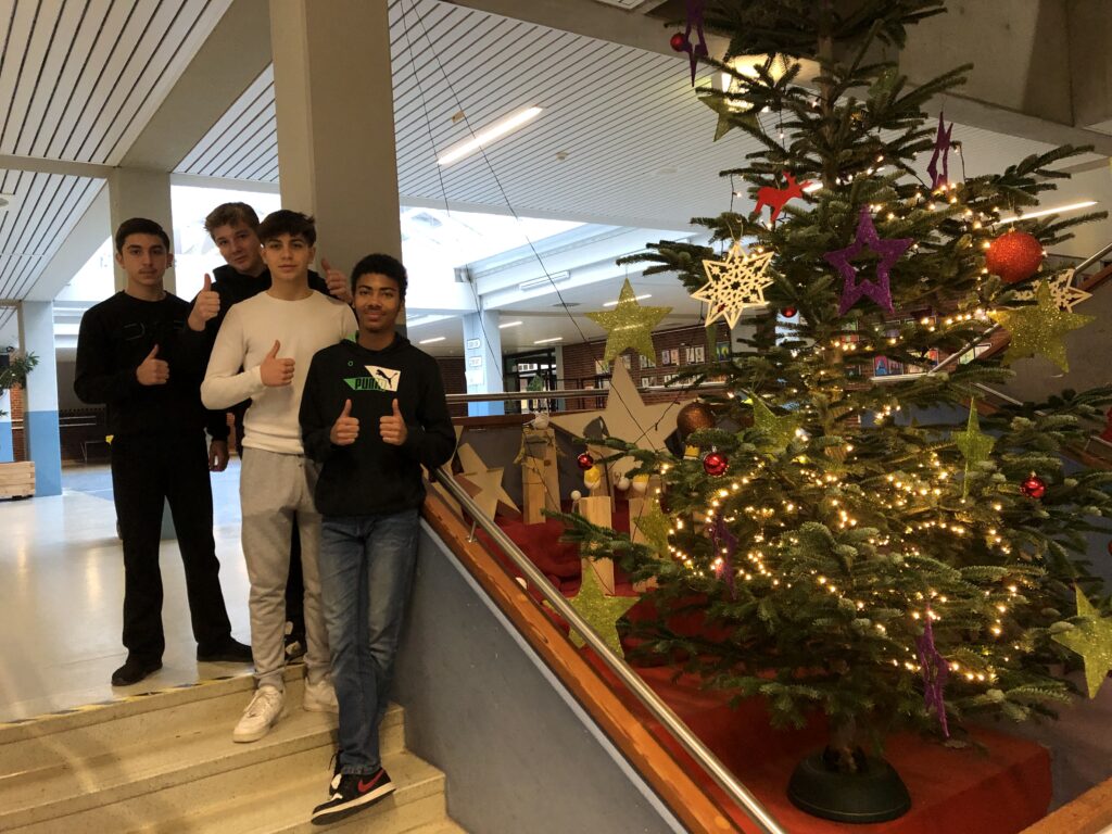 Das Bild zeigt den geschmückten Weihnachtsbaum im Foyer der Schule und links daneben stehend 3 Schüler