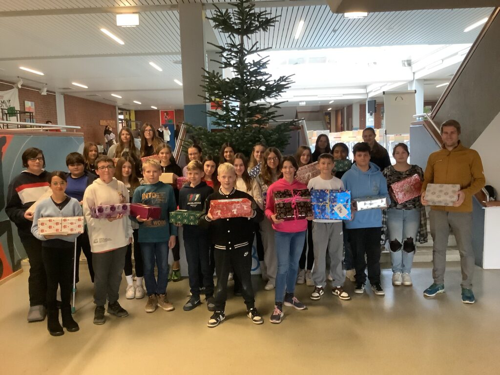 Lehrer Markus Hantke steht mit seiner Klasse im Foyer vor einem Weihnachtsbaum und alle Kinder halten Päckchen mit Geschenken für bedürftige Kinder