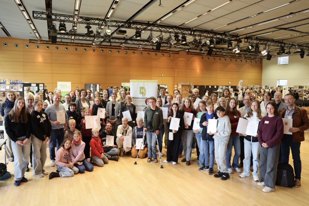 Vertreter/innen der 21 geehrten Umweltschule präsentieren in einem Halbkreis ihre Urkunden