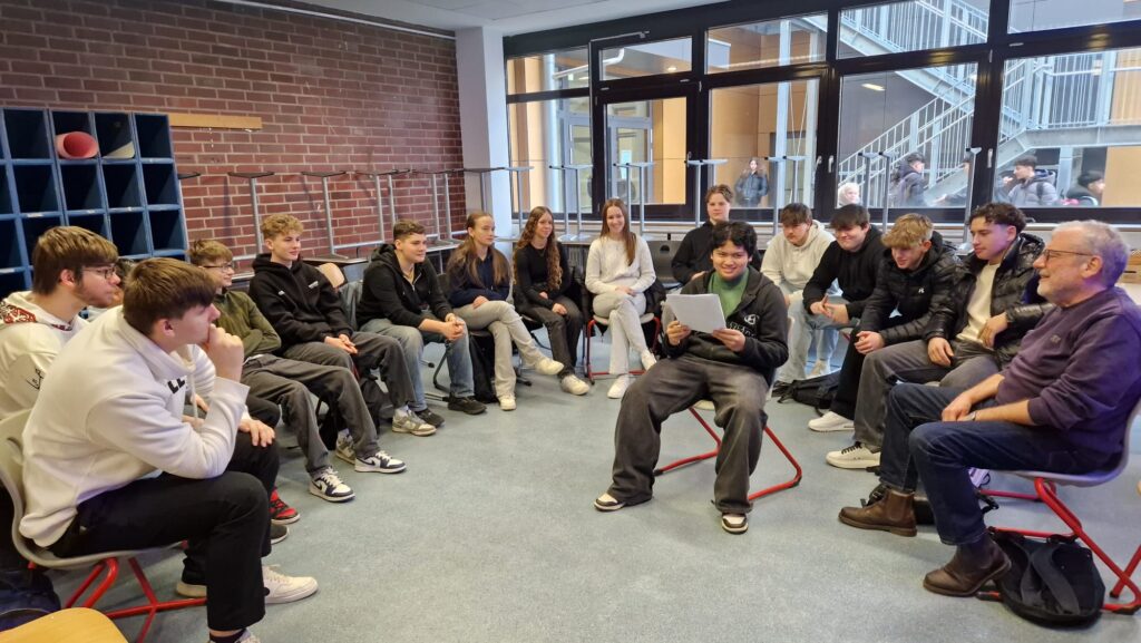 Schüler/innen sitzen im Stuhlkreis um einen Schüler, der in der Mitte sitzt und etwas vorliest