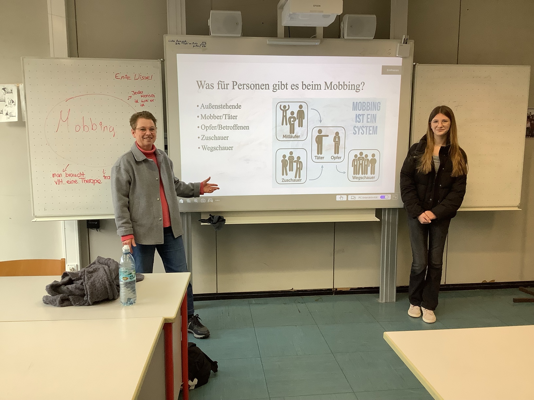 Frau Holzapfel-Hesselmann steht links neben dem Smartboard zum Thema Mobbing und eine Schülerin steht rechts neben dem Smartboard
